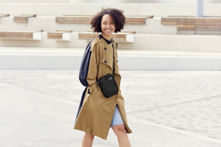 A woman models the Bellroy City Pouch, worn crossbody over a tan trench coat and denim dress in a modern urban outdoor setting.