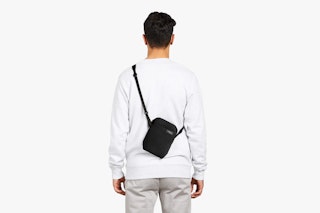 The model wears a light gray looma_weave sweatshirt and trousers and presents the black Bellroy City Pouch crossbody bag on a white background.
