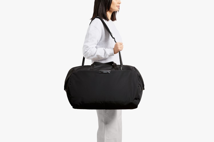A person holds the Bellroy Classic Weekender 45L duffel, showing black recycled ripstop fabric and dual straps on a white backdrop.