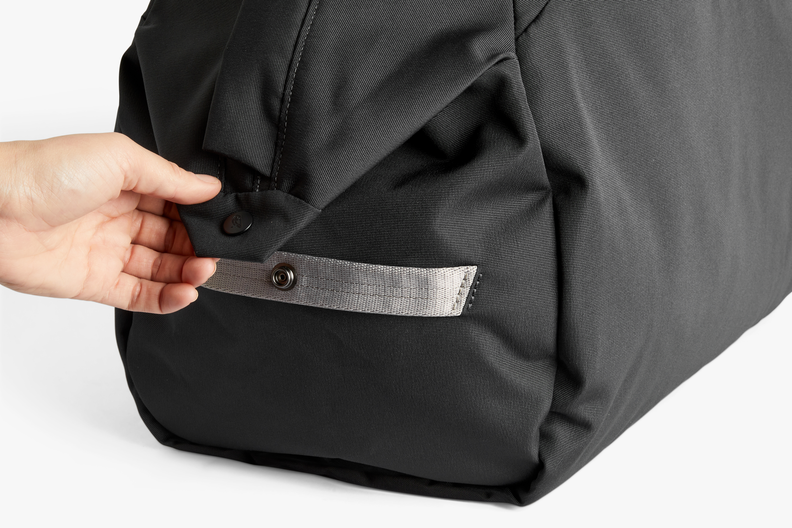 A hand opening a press stud on the corner of the black Bellroy Classic Weekender 45L reveals a strap and textured fabric detailing.