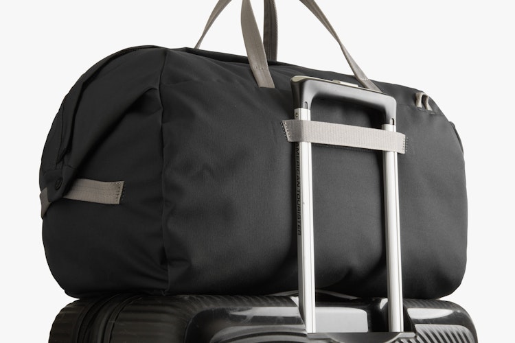 Bellroy Classic Weekender 45L in black ribba weave with beige handles, secured atop a slate suitcase, shown on white background.