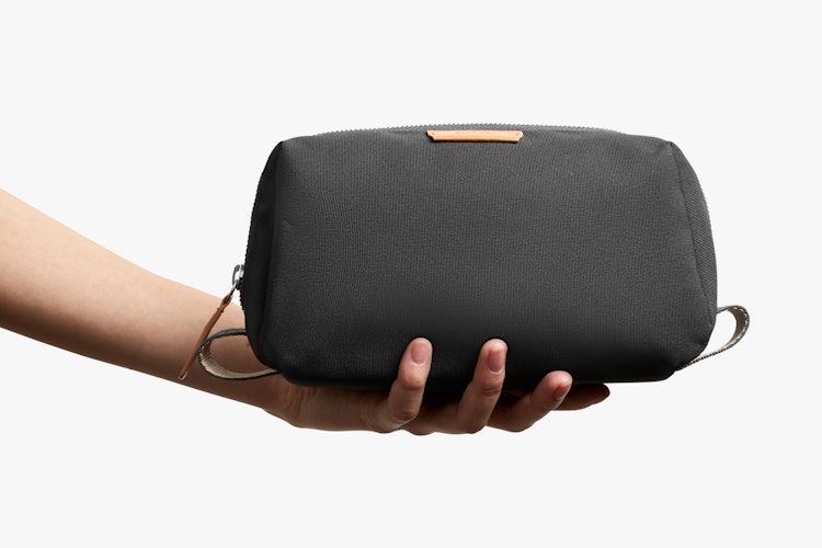 One hand shows the Bellroy Toiletry Kit, a black case with zipper, ribba fabric and brown leather accents on a white background.