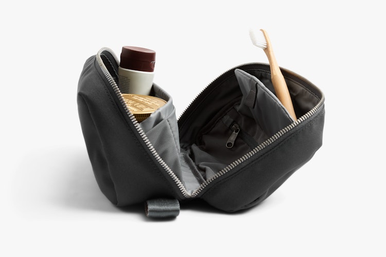 Bellroy Toiletry Kit Plus in slate-colored Ribba fabric, opened, with a wooden toothbrush, a small bottle and a tin on a white background.