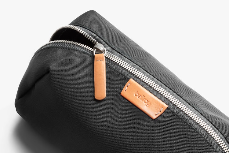 Bellroy Toiletry Kit Plus in black with ribbed fabric, silver zipper, tan leather pull tab, and Bellroy patch shown in detail.