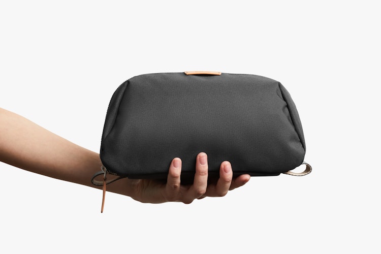 One hand displays the Bellroy Toiletry Kit Plus, a slate-colored rectangular pouch with a tan zipper and textured fabric, on a white background.