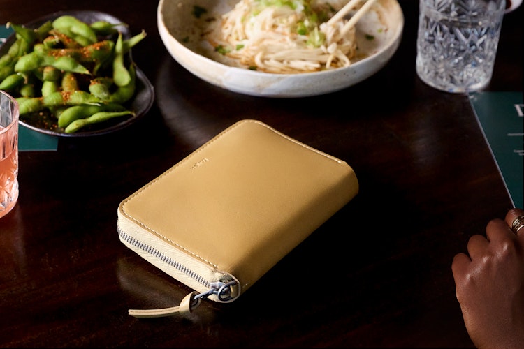 Beige Bellroy Folio Wallet with RFID protection on a dark wooden table, shown in use next to food, drinks, and a hand with rings.