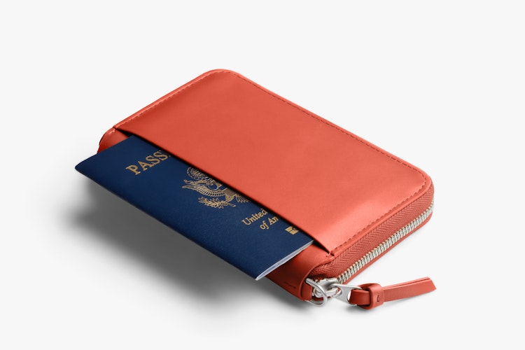 Bellroy Folio Wallet in blood orange with RFID protection, closed and with zipper, shows a passport in the outer pocket on a white background.