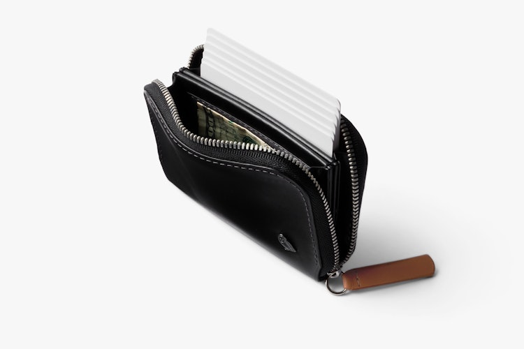 The Bellroy Folio Mini, a black leather RFID wallet with zipper, is shown on a white surface with coins, blank cards and folded cash.